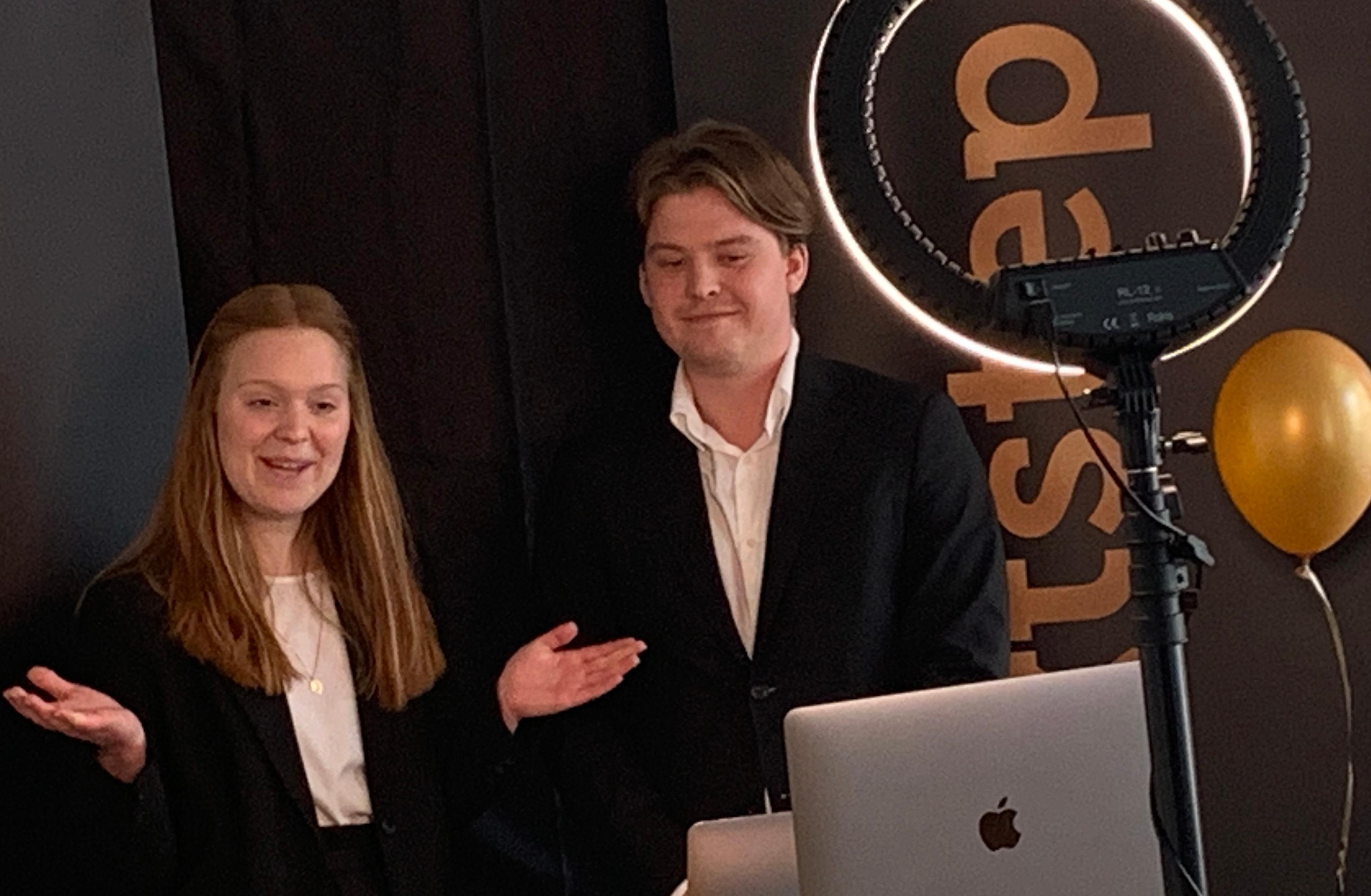 Two students stand in front of a laptop, filming themselves in a room with a black backdrop. 