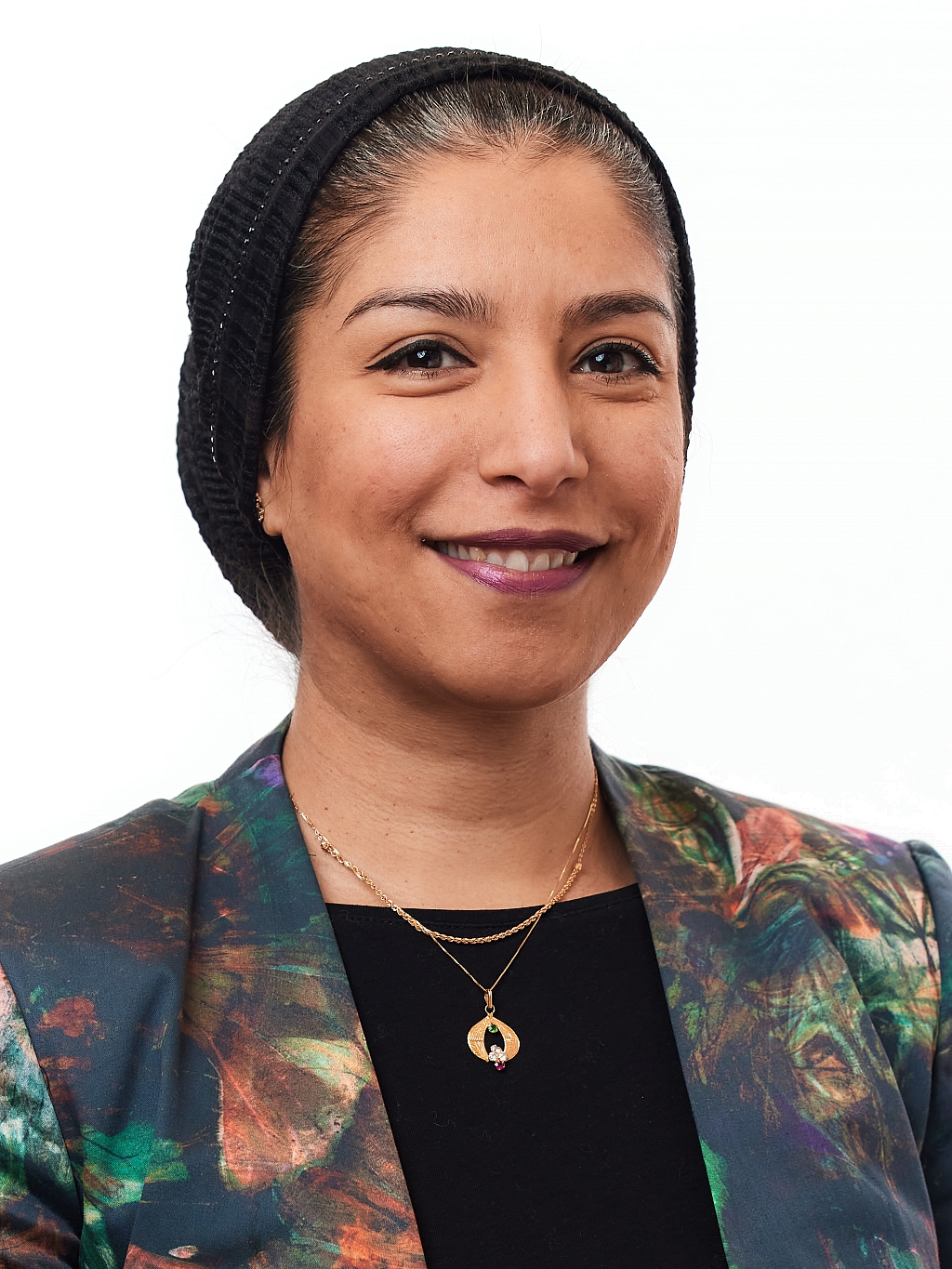 Woman wearing a dark headscarf, patterned blazer, and gold necklaces, smiling and facing forward against a white background.
