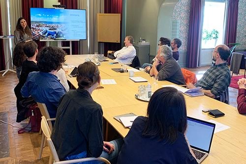 Meeting room with a long table and a large screen at one end. Researchers are sitting around the table and one is standing up giving a presentation. 