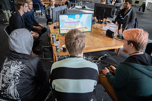 Image shows a large room with young tables and computers. A group of young men sit around a table that has computer monitors on it. They are playing a video game. 