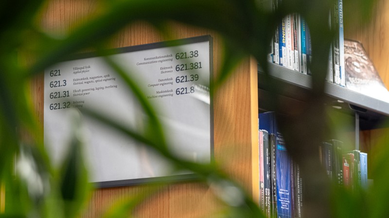 A shelf end with a sign showing the shelf numbers for the shelf. The image is taken through a plant that is partially out of focus but frames the picture.