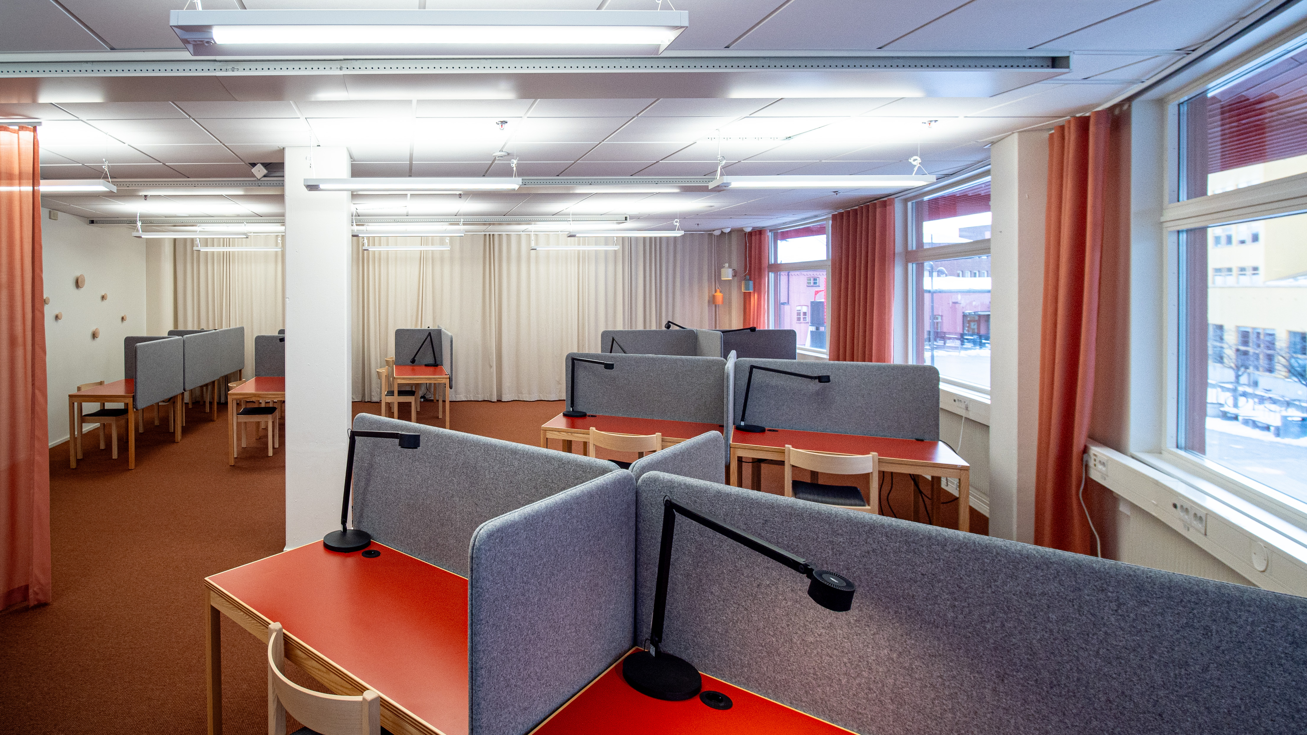 A room with several tables displayed with screens that separate the tables from each other. The room’s floor is covered with an orange wall-to-wall carpet, and large windows are visible to the right in the picture.