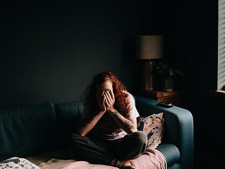 Girl sits on bed in darkened room, her face is in her hands. 