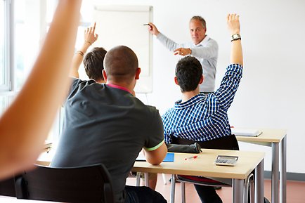 Students raising their hands in the foreground, teacher in the background